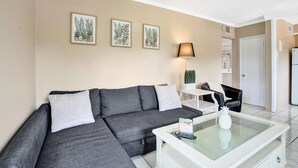 Ein modernes Wohnzimmer mit einem grauen Ecksofa, einem weißen Couchtisch und einem kleinen weißen Tisch mit einer Lampe. An der Wand hängen drei gerahmte botanische Drucke.