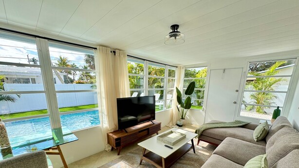 A living room with a TV, sofa, coffee table, and large windows overlooking a pool. - Beautiful 3 2 Home in Pompano Beach - Close to Beach (Pompano Beach)