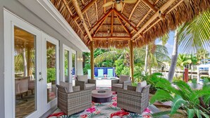 A covered outdoor seating area with wicker furniture and a thatched roof, surrounded by lush greenery. - Flirty Flamingo Colorful Waterfront Escape w Pool (Wilton Manors)