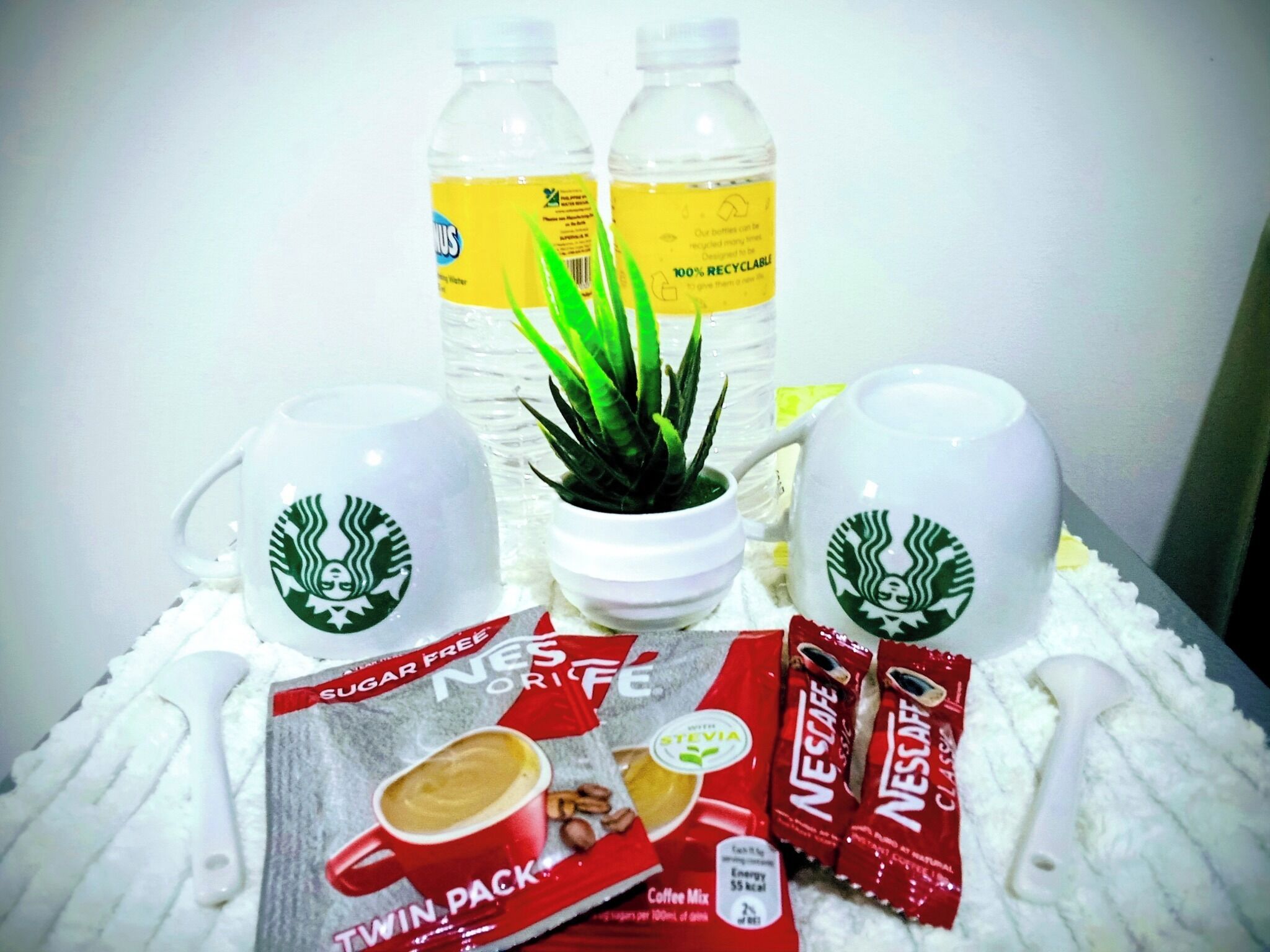 Ein Tisch mit zwei Starbucks-Tassen, einer Topfpflanze, zwei Wasserflaschen und einer Packung Nescafe-Kaffee.