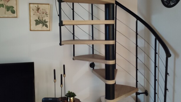 Un escalier en colimaçon, avec des marches en bois et une rampe en métal noir, menant à l’étage supérieur.