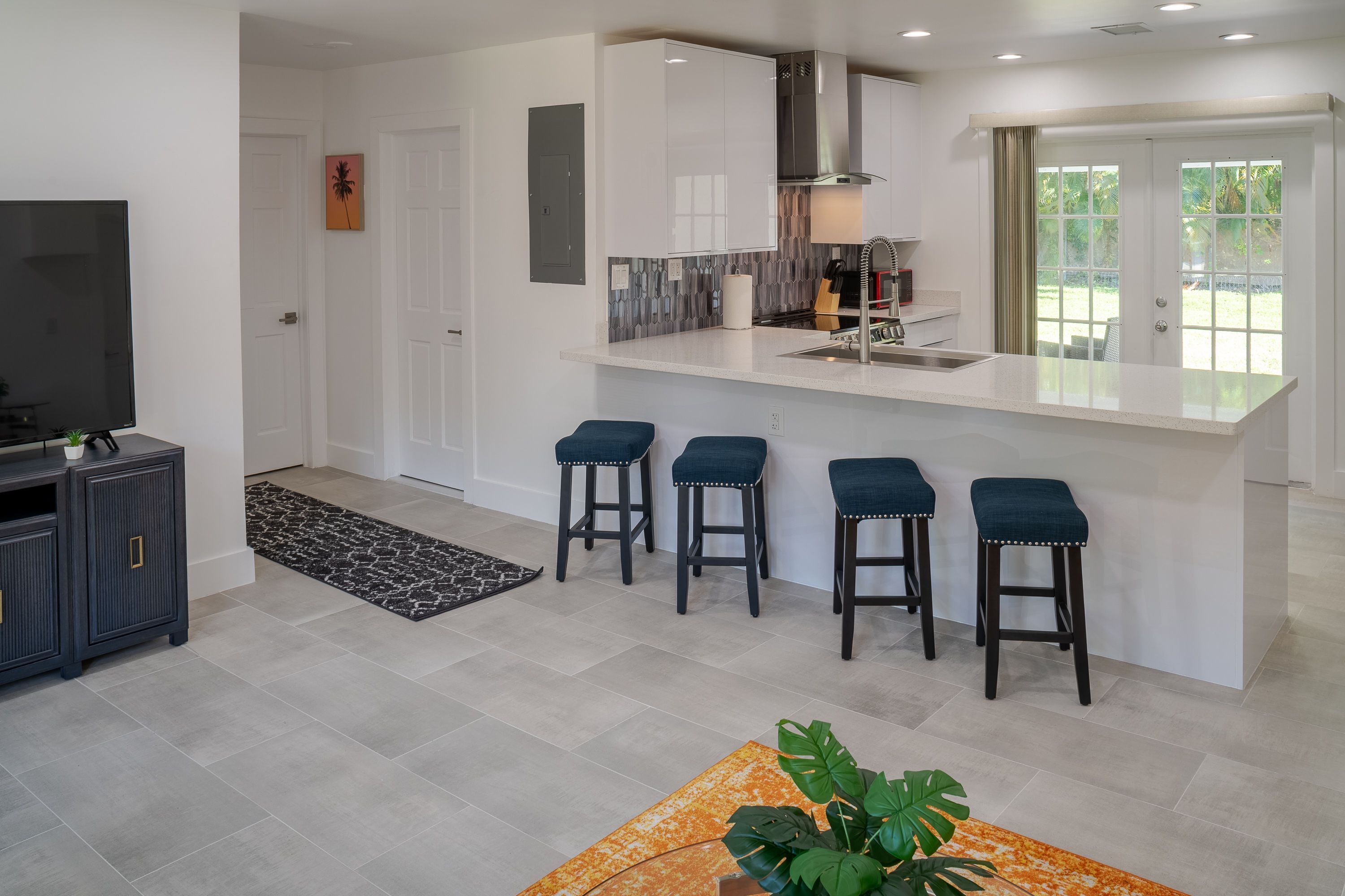 A modern kitchen with a central island, bar stools, a TV, and a dining area with a patterned rug.