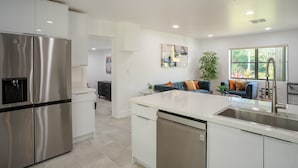 A modern kitchen with stainless steel appliances, a large island with a sink, and a view of a living area with a sofa and plants. - Spacious 3BR Home 2 Baths Amazing Pool Area (Wilton Manors)