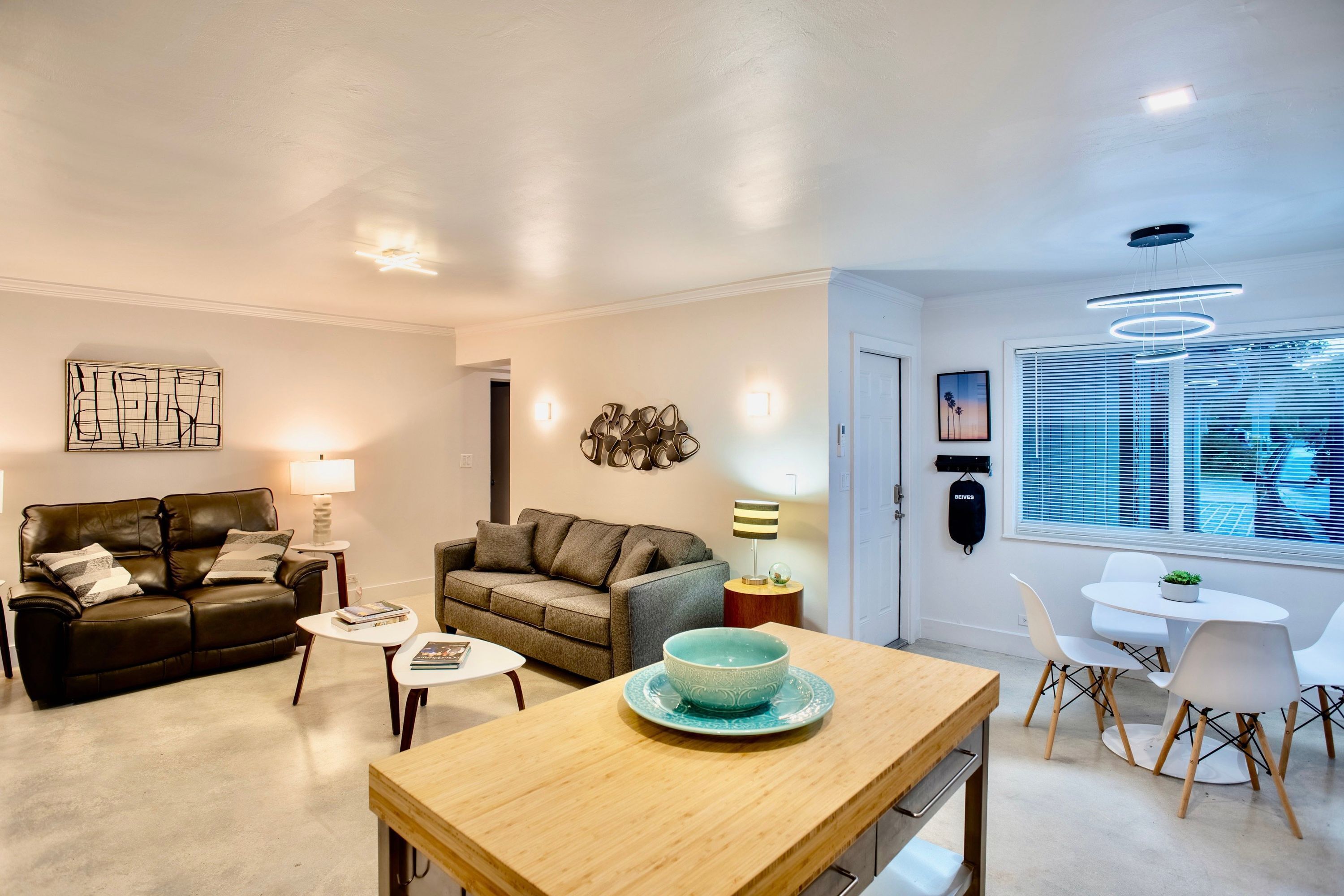 Ein modernes Wohnzimmer mit einem braunen Ledersofa, einem grauen Sofa, einem Holzespeisetisch mit Stühlen und einem großen Fenster mit Jalousien.