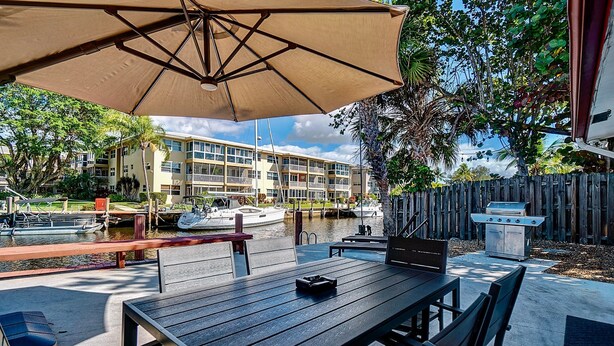 An outdoor patio with a table and chairs, a grill, and a view of a marina with boats and a building in the background. - Citrus Isles Waterfront Home (Fort Lauderdale)