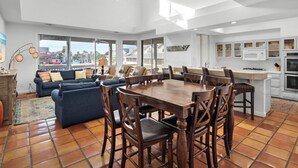 A spacious living area with a large dining table, comfortable seating, and large windows offering views of the city.