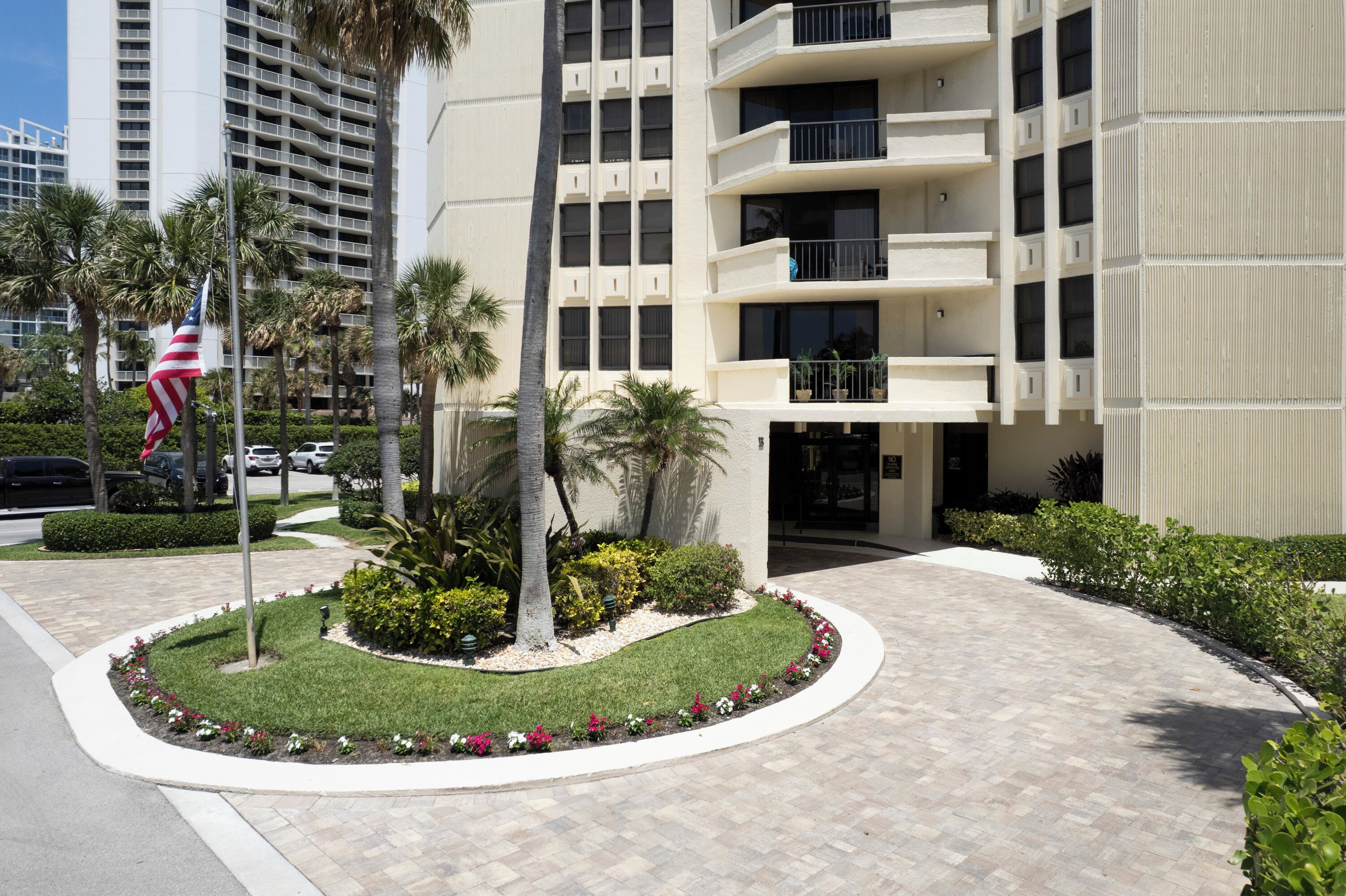 A modern building with a landscaped entrance featuring a flag, palm trees, and a well-maintained garden.