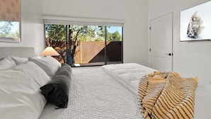 Une chambre à coucher avec un lit, une fenêtre avec des stores, un téléviseur fixé au mur et une vue sur l’extérieur.