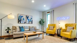 Ein modernes Wohnzimmer mit einer Couch, zwei Sesseln, einem Couchtisch und einer Stehlampe. An der Wand befinden sich abstrakte Gemälde und ein Neon-Schild.
