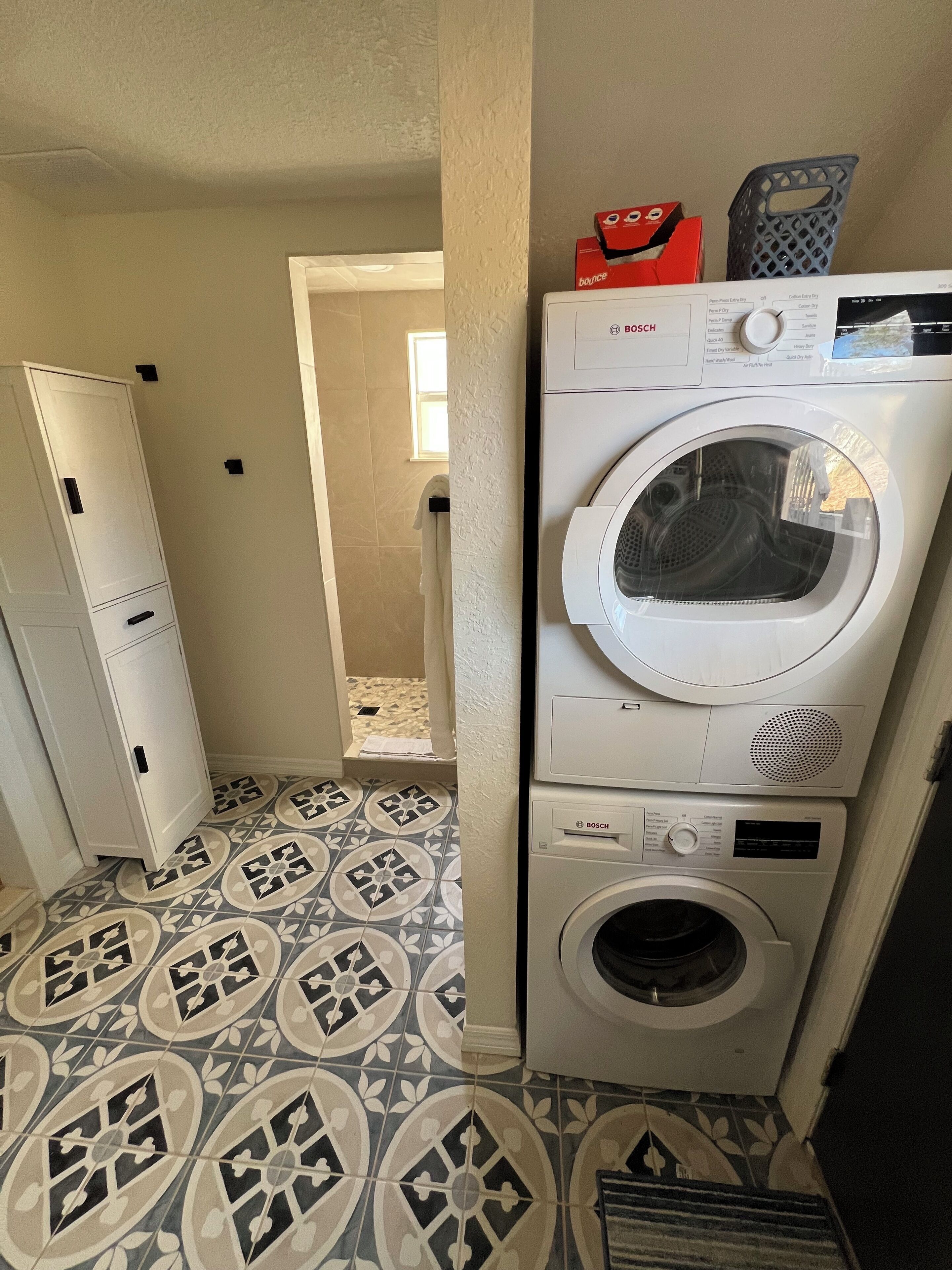 Un espace buanderie équipé d’un lave-linge et d’un sèche-linge Bosch, de carreaux de sol à motifs et d’un meuble blanc.