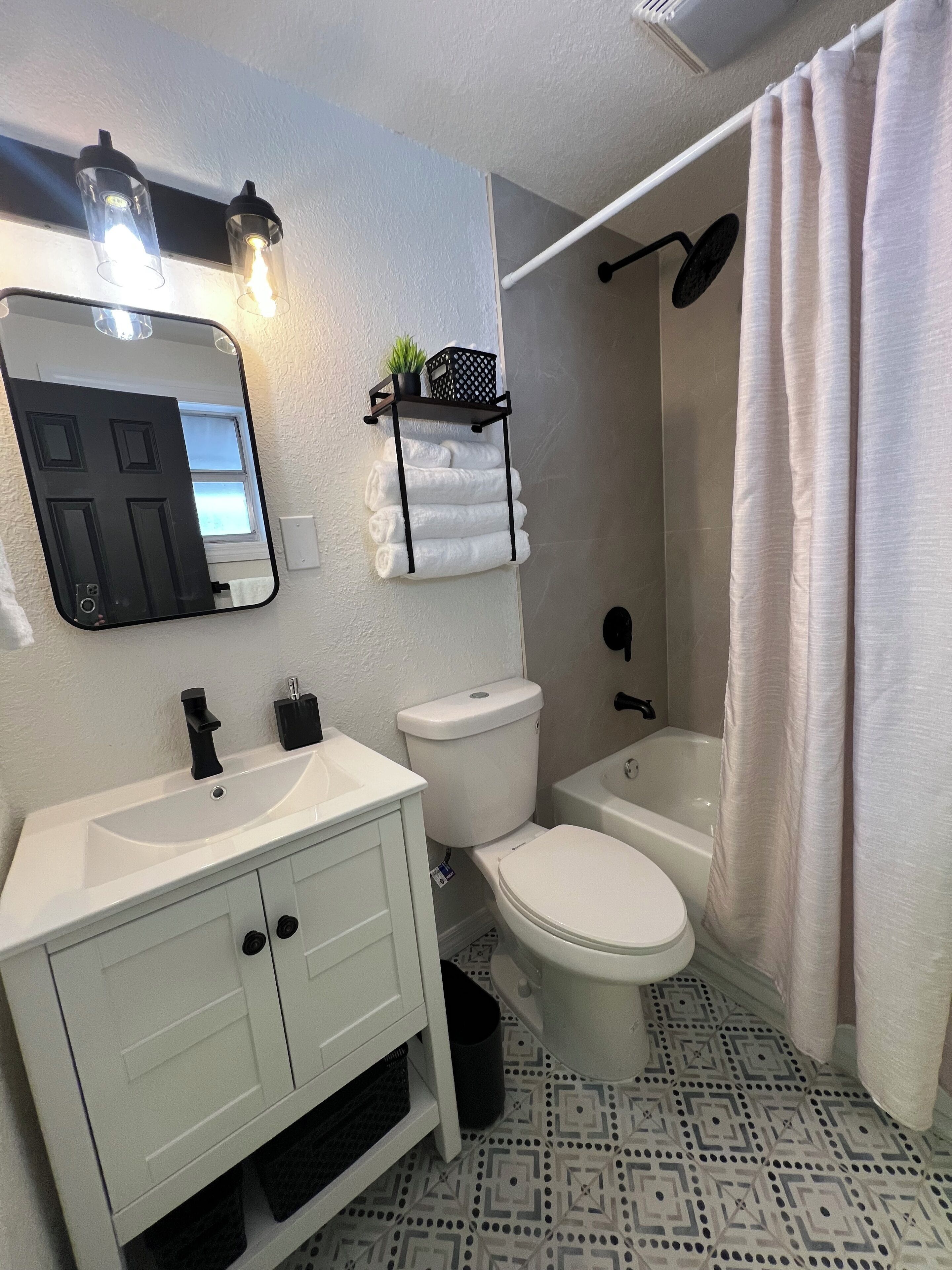 A bathroom with a white sink, toilet, and bathtub. There is a towel rack with folded towels and a small plant on it. The floor has a geometric tile pattern.