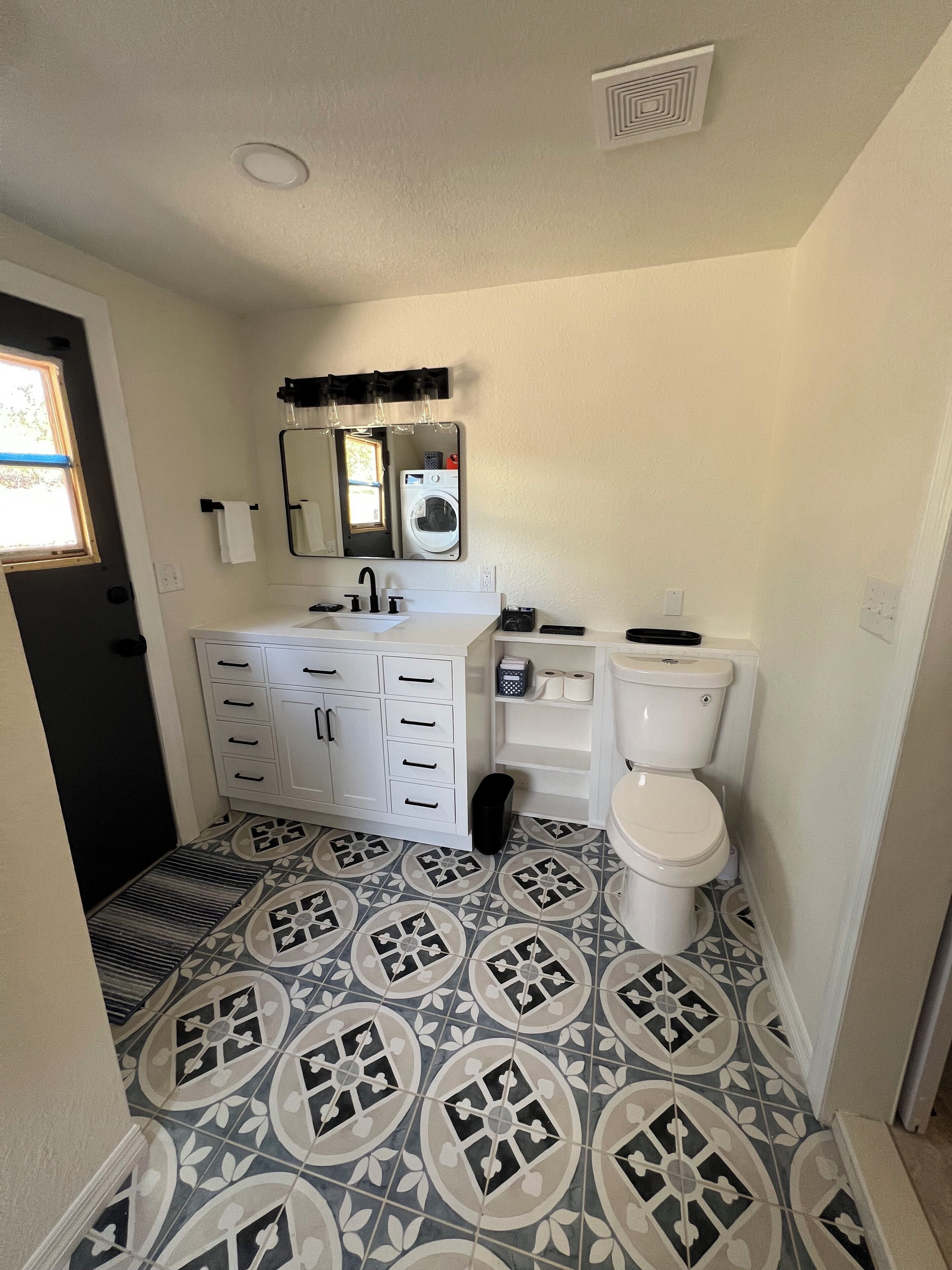 A bathroom with a patterned tile floor, a white sink with a mirror above it, a toilet, and a black trash bin.
