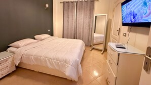 Un dormitorio con una cama, una mesita de noche, un tocador, un espejo y un televisor en la pared.