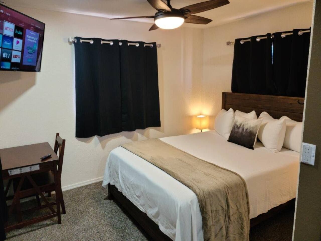A bedroom with a bed, a desk, a chair, a ceiling fan, and a TV mounted on the wall.