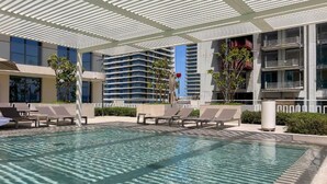 A pool area with lounge chairs, a pergola, and a modern building in the background.
