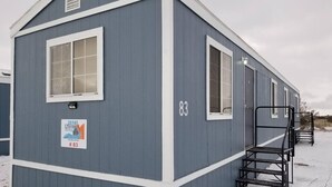 A small, single-story building with a blue exterior and white trim. It has a sign indicating it is a Grand Canyon retreat, number 83. There is a metal staircase leading to a door on the right side.