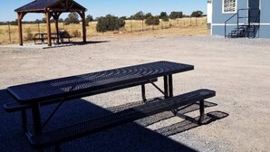 A picnic table with a bench in a dry, open field with a small wooden structure and a building in the background.