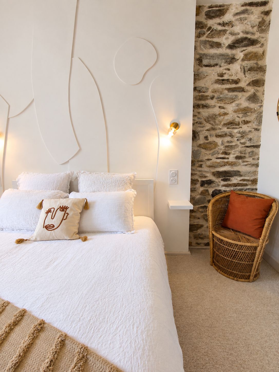 Een slaapkamer met een groot bed, witte beddengoed, een accentmuur van steen, een rieten stoel en een wandlamp.