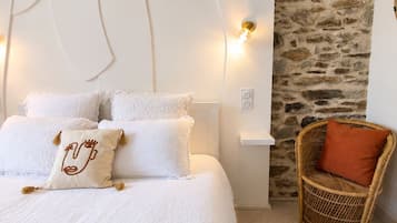Een slaapkamer met een groot bed, witte beddengoed, een accentmuur van steen, een rieten stoel en een wandlamp.