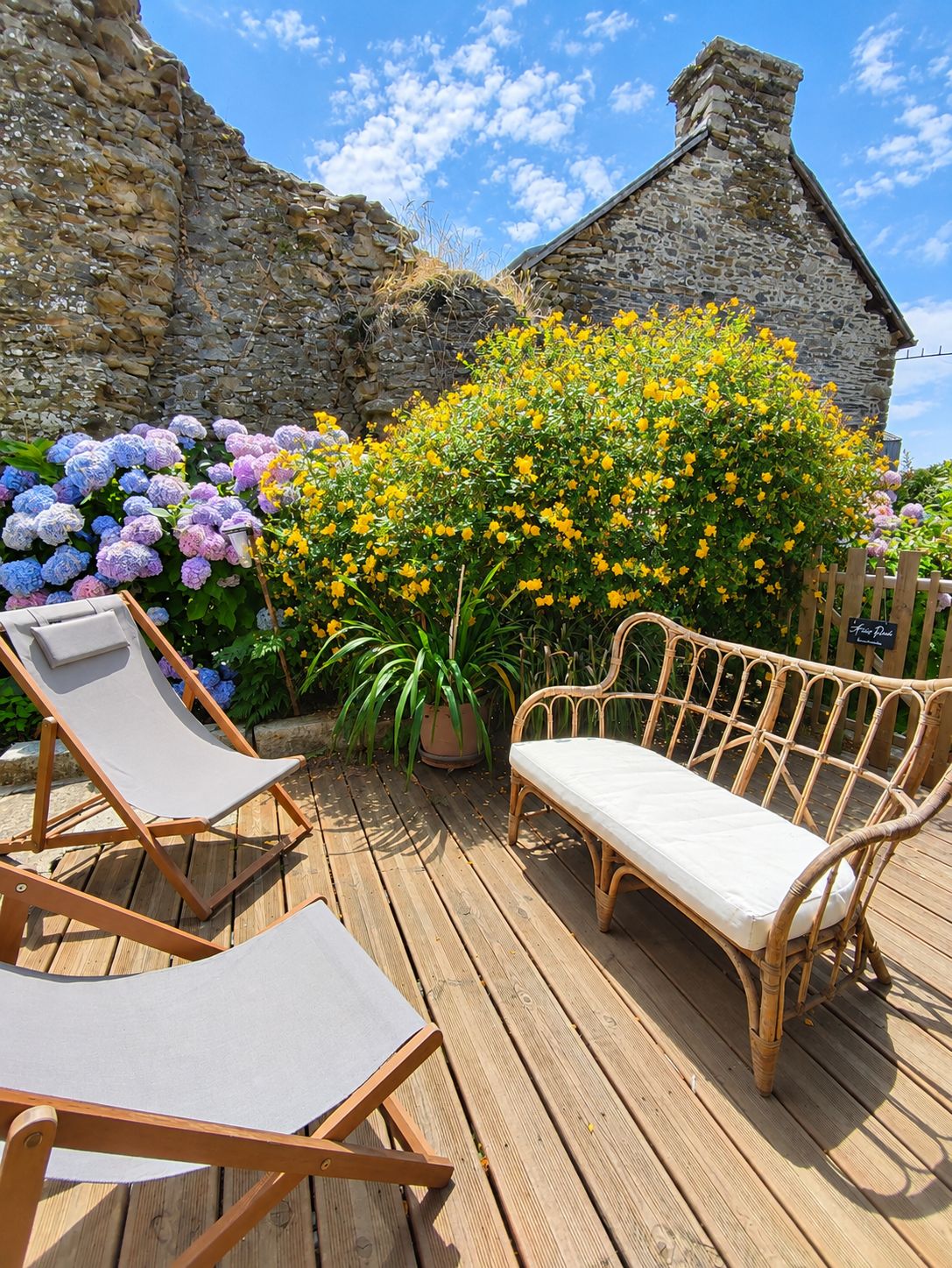 Een houten terras met twee lounge stoelen en een rieten bankje, omgeven door diverse bloeiende planten en een stenen muur.