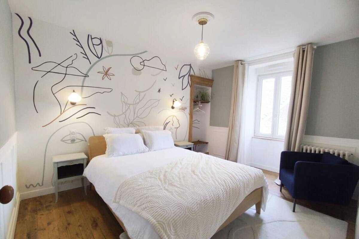 A bedroom with a large bed, a bedside table, a chair, a wall with abstract art, and a window with curtains.