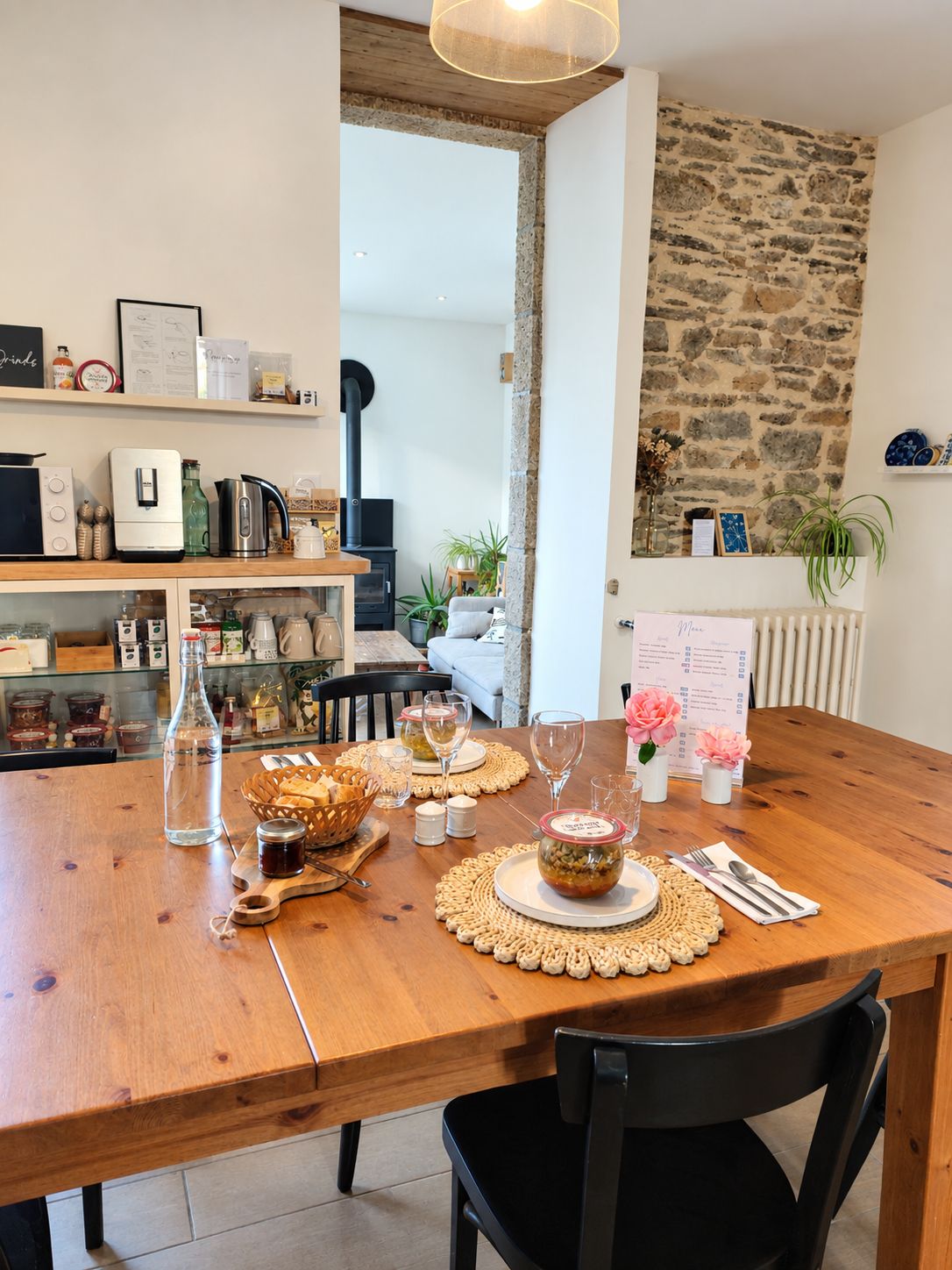 A dining area with a wooden table set for two, a stone wall, and a kitchen area with various appliances and items on the shelves.