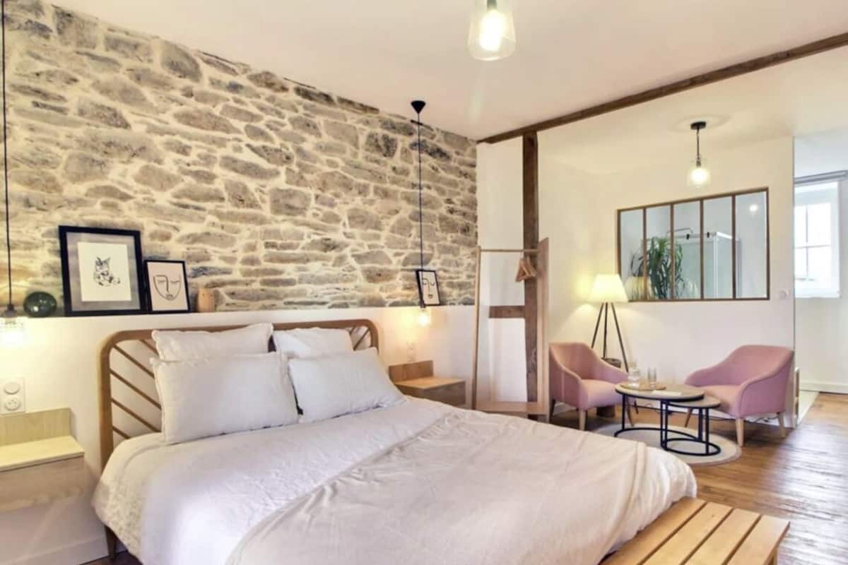 A bedroom with a stone wall, a bed with white bedding, a wooden headboard, a nightstand, a small table with a lamp, and two chairs.