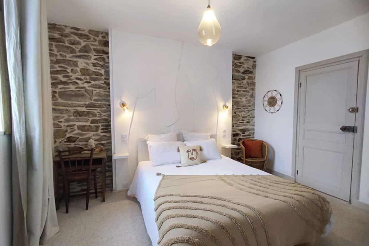 A bedroom with a stone wall, a bed with a beige blanket, a wooden chair, a small table, and a door.