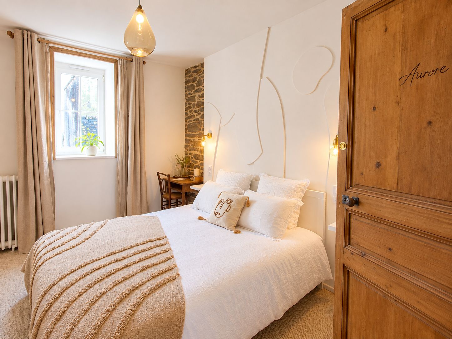 Un dormitorio con una cama, un escritorio, una silla, un ventanal con cortinas, una pared de piedra y un armario de madera.