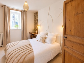 A bedroom with a bed, a desk, a chair, a window with curtains, a stone wall, and a wooden wardrobe. - Les Chambres D'Hespéris (Pleyben)