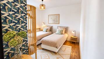 Un dormitorio con dos camas, un panel de madera, una pared con un diseño de hojas, una obra de arte enmarcada y una mesita con un jarrón.