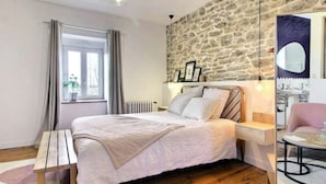 Un dormitorio con pared de piedra, una cama con cabecera, una mesita de noche, una silla y un ventanal con cortinas.