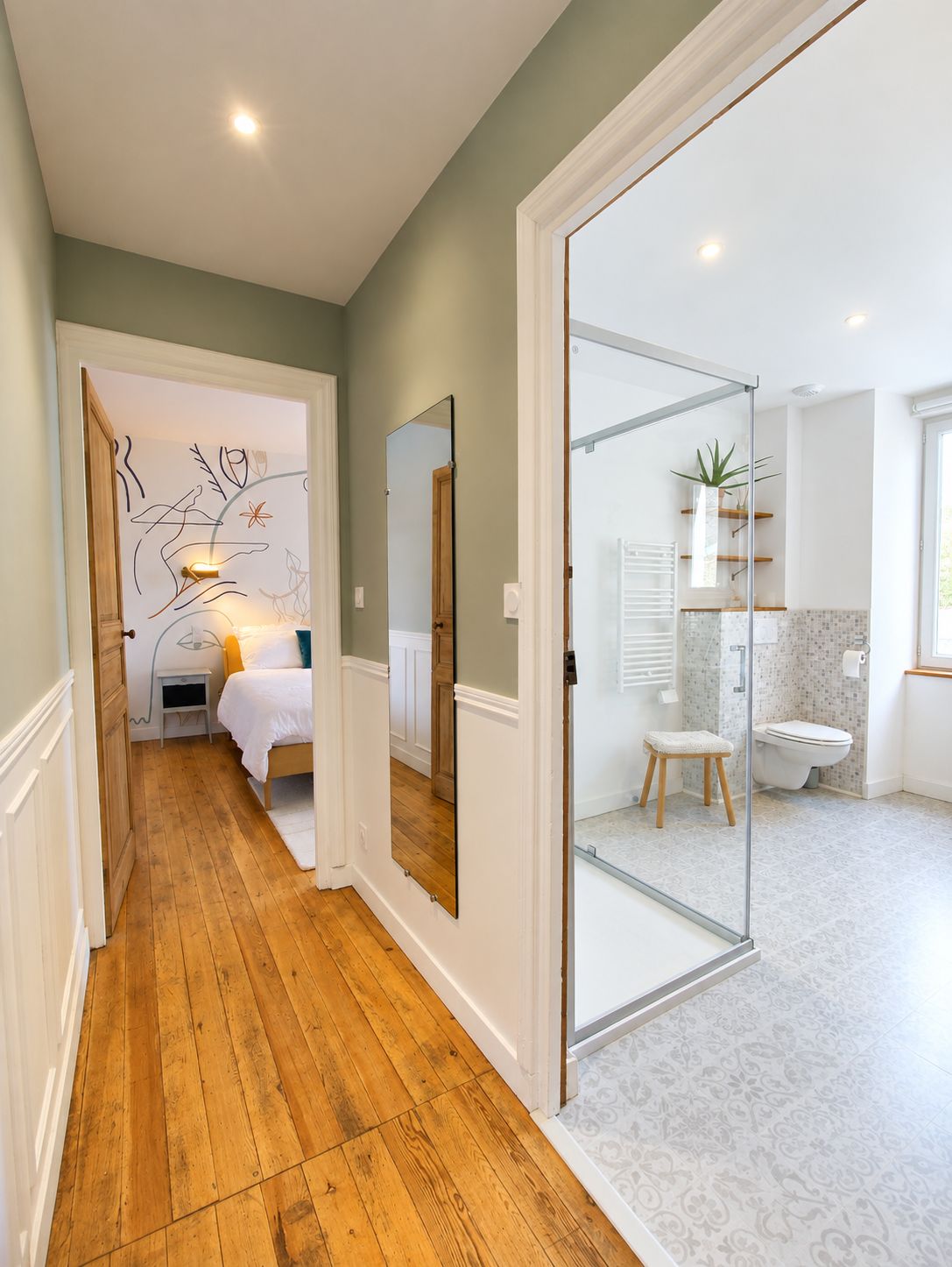 Een gang die uitkomt in een slaapkamer met houten vloer, een bed en een spiegel aan de muur.