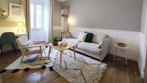 Una sala de estar con un sofá, una silla, una mesa de centro y una mesita auxiliar. Hay una ventana con cortinas, un radiador y una lámpara colgante.