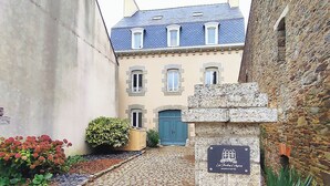 Un estrecho callejón adoquinado con una puerta azul, una pared de piedra con una placa conmemorativa, y un edificio con numerosas ventanas y tejado de tejas.