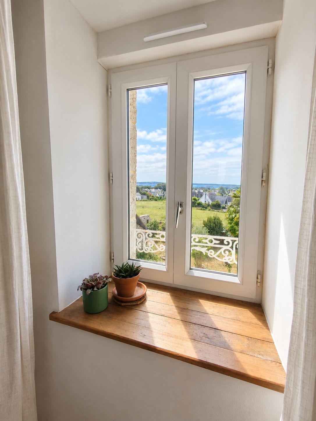 Een kamer met een raam dat uitkijkt op een balkon met uitzicht op groen en gebouwen. Op de vensterbank staan twee potplanten.