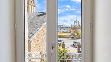 Une fenêtre donnant sur un bâtiment historique doté d’un clocher, une rue animée par la circulation et un espace jardin.