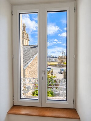A window with a view of a historic building with a bell tower, a street with cars, and a garden area. - Les Chambres D'Hespéris (Pleyben)