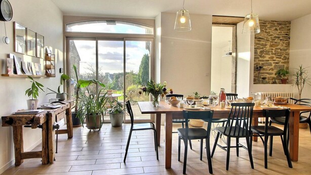 Un comedor con una mesa grande, sillas y vistas al exterior. Hay plantas en macetas y un reloj en la pared.