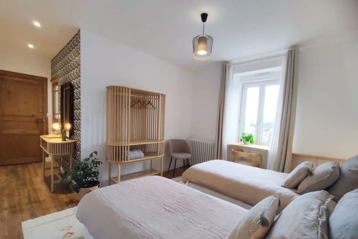 A bedroom with two beds, a wooden wardrobe, a desk, a chair, and a hanging plant.