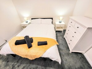 Un lit bien fait, avec une couette blanche, deux serviettes noires et un plaid jaune. Il y a deux tables de chevet avec des lampes, une commode blanche avec de nombreux tiroirs et un sol recouvert d’une moquette grise.