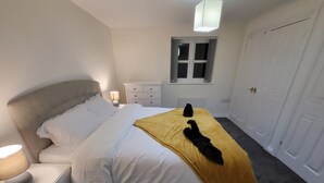 Una cama bien hecha con edredón blanco y manta amarilla, una lámpara de noche, una mesita de noche blanca, una ventana pequeña con vistas y una puerta blanca.