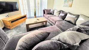Una sala de estar con un televisor de pantalla plana grande, una mesa de centro de madera, un sofá gris con varios cojines y una ventana con persianas.