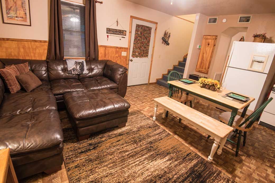 A cozy living room with a brown leather sofa, a wooden dining table with a bench, and a kitchen area with a refrigerator and a wooden cabinet.