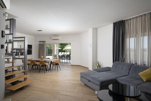 Een moderne woonkamer met een grijze bank, een houten trap, een eethoek en grote ramen.