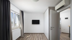 Una habitación con piso de madera, una puerta blanca, un televisor negro montado en la pared y un ventanal con cortinas.
