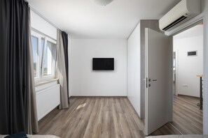 Una habitación con piso de madera, una puerta blanca, un televisor negro montado en la pared y un ventanal con cortinas.