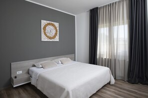Un dormitorio moderno con una cama, una mesita de noche, un enchufe en la pared y un ventanal con cortinas translúcidas.
