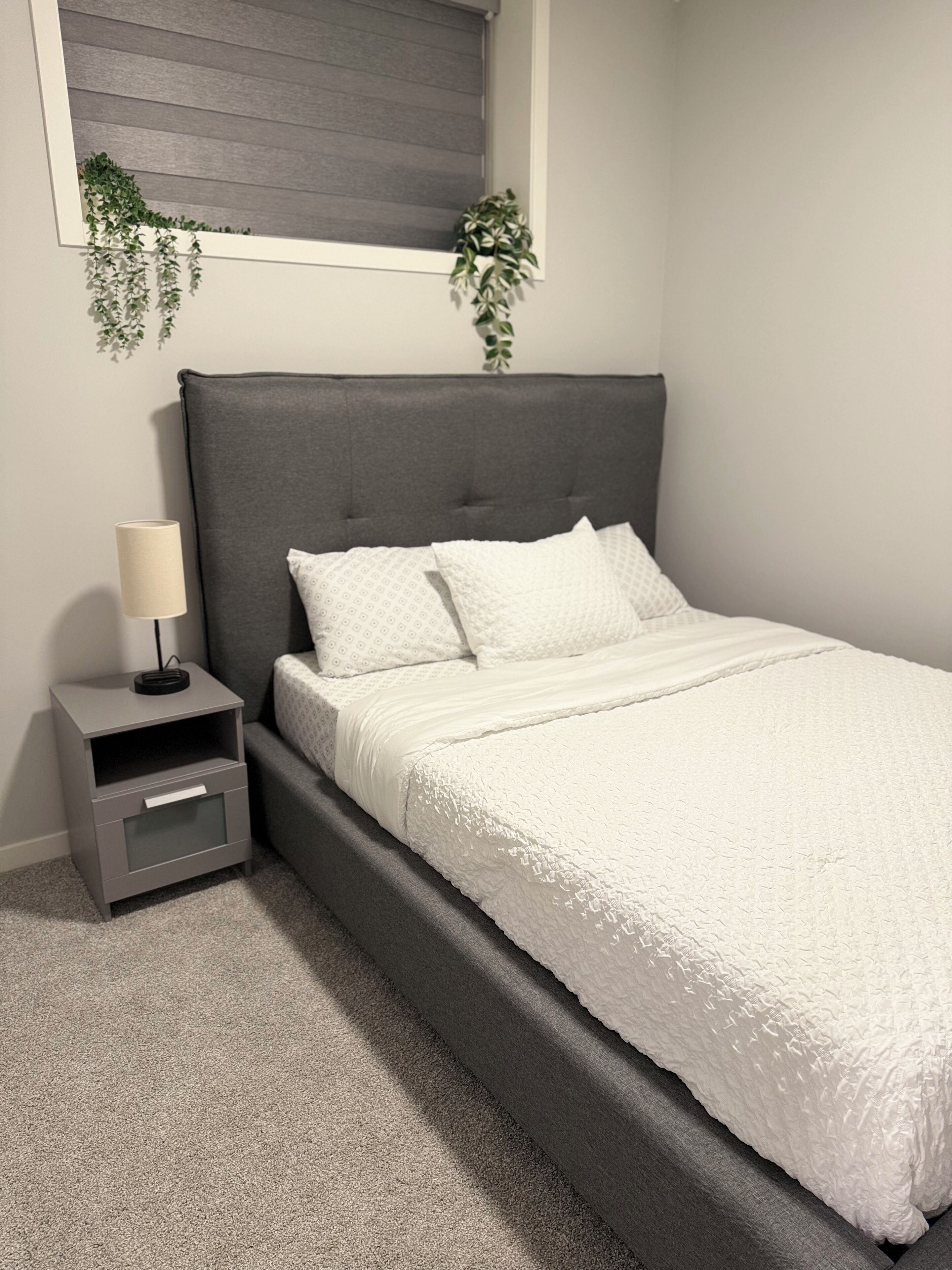 A bedroom with a grey upholstered headboard, a white quilted bedspread, a bedside table with a lamp, and a small plant on the windowsill.