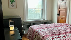 Ein Schlafzimmer mit einem Bett, einem Schreibtisch mit Lampe, einer Heizung, einem Fenster mit Blick auf Gebäude und einer Tür.